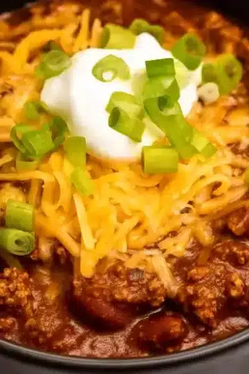 Loaded crock pot chili with ground beef and kidney beans served in black bowl, generously topped with shredded cheddar cheese, sour cream dollop, and fresh sliced green onions on white marble countertop