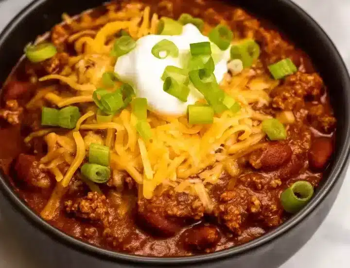 Loaded crock pot chili with ground beef and kidney beans served in black bowl, generously topped with shredded cheddar cheese, sour cream dollop, and fresh sliced green onions on white marble countertop