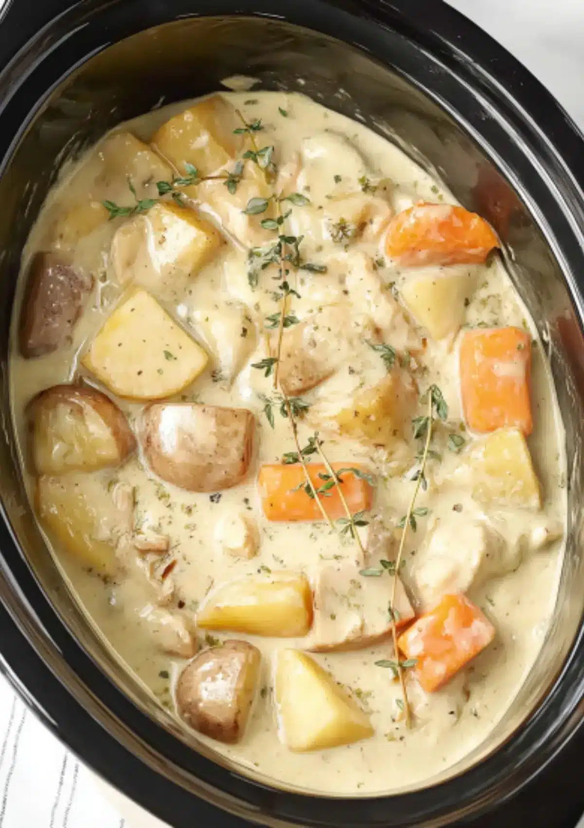 Overhead view of creamy ranch chicken and potatoes in black crock pot slow cooker with chunks of chicken, potatoes, orange carrots and fresh thyme garnish in rich cream sauce