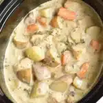 Top view of slow cooker creamy ranch chicken and potatoes recipe with visible potato chunks, baby carrots, tender chicken pieces in creamy beige sauce, garnished with fresh thyme sprigs