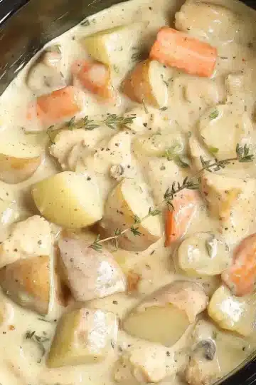 Top view of slow cooker creamy ranch chicken and potatoes recipe with visible potato chunks, baby carrots, tender chicken pieces in creamy beige sauce, garnished with fresh thyme sprigs