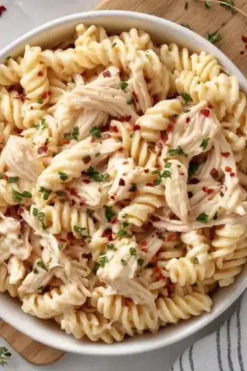 Overhead view of creamy garlic parmesan chicken pasta in a white bowl positioned on a wooden cutting board. Fresh thyme sprigs are scattered around the board, and a gray and white striped kitchen towel is placed in the bottom right. The rotini pasta is mixed with shredded chicken and garnished with parsley and red pepper flakes on a white marble surface