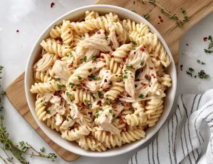 Overhead view of creamy garlic parmesan chicken pasta in a white bowl positioned on a wooden cutting board. Fresh thyme sprigs are scattered around the board, and a gray and white striped kitchen towel is placed in the bottom right. The rotini pasta is mixed with shredded chicken and garnished with parsley and red pepper flakes on a white marble surface