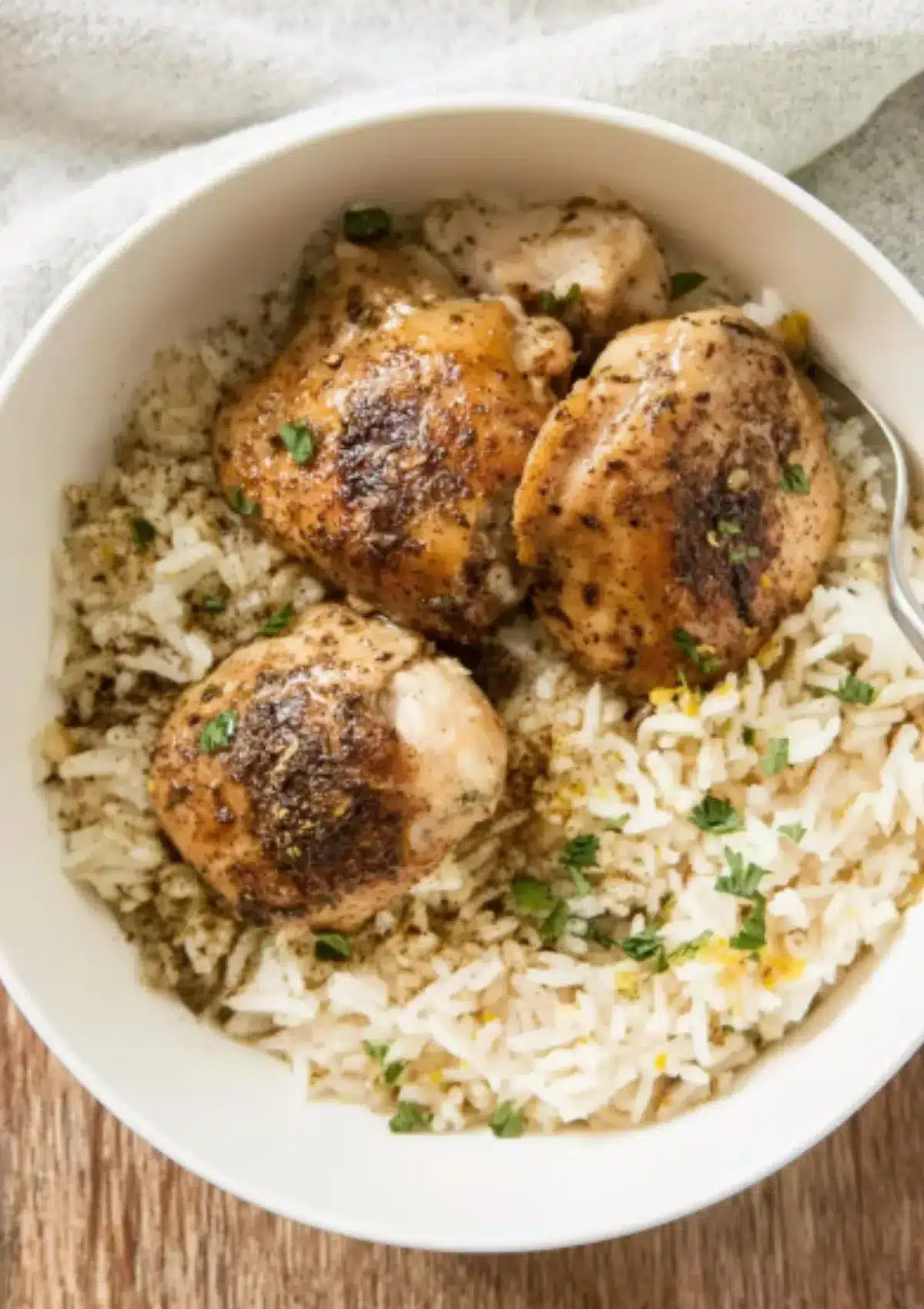 Overhead view of three golden-brown boneless chicken thighs with paprika and Italian herb seasoning served over fluffy jasmine rice in white ceramic bowl, garnished with fresh parsley, on wooden table with linen napkin