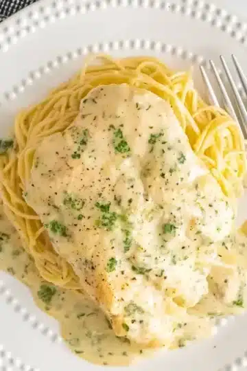 Overhead view of Crock Pot Angel Chicken served on decorative white plate with dotted edges, tender chicken breast in rich cream sauce over perfectly cooked angel hair pasta, garnished with fresh parsley and black pepper, silver fork placed on right side, black and white checkered napkin in corner