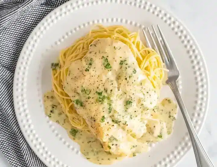 Overhead view of Crock Pot Angel Chicken served on decorative white plate with dotted edges, tender chicken breast in rich cream sauce over perfectly cooked angel hair pasta, garnished with fresh parsley and black pepper, silver fork placed on right side, black and white checkered napkin in corner