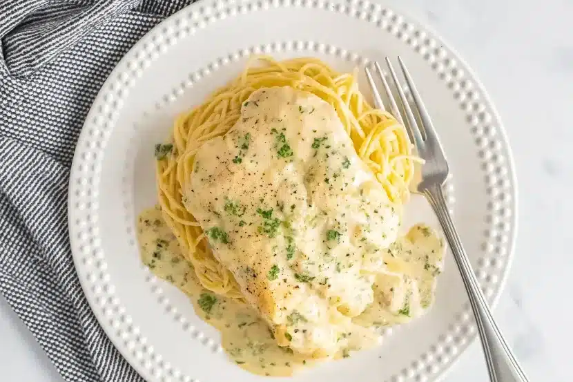 Overhead view of Crock Pot Angel Chicken served on decorative white plate with dotted edges, tender chicken breast in rich cream sauce over perfectly cooked angel hair pasta, garnished with fresh parsley and black pepper, silver fork placed on right side, black and white checkered napkin in corner