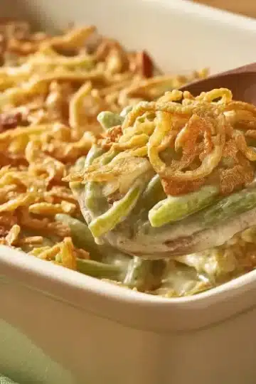 Close-up of wooden spoon holding portion of green bean casserole showing creamy texture and crispy fried onion topping over white baking dish