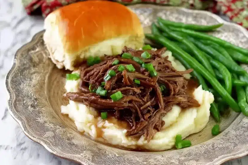 Fork-tender Mississippi pot roast shredded over creamy mashed potatoes, garnished with green onions, served with green beans and buttery dinner roll on ornate plate against white marble background