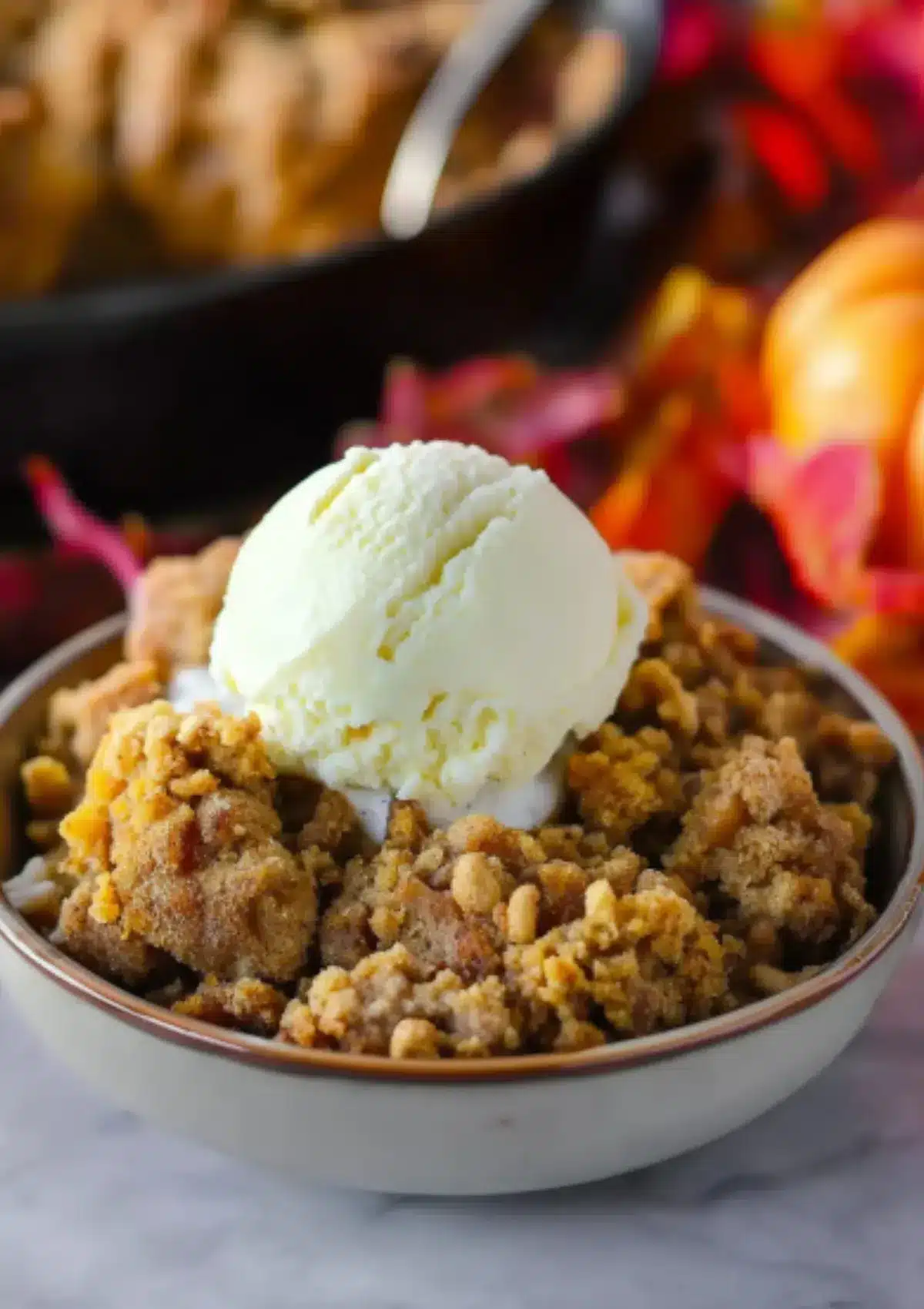 Pumpkin crisp dessert in white bowl with cream-colored rim, topped with vanilla ice cream scoop, golden cinnamon streusel visible, black cast iron skillet and autumn leaves in background