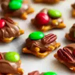 Overhead view of multiple Rolo pretzel bites arranged in neat rows on white marble surface, featuring variety of toppings including red and green M&Ms with white "m" letters and brown pecan halves on melted chocolate