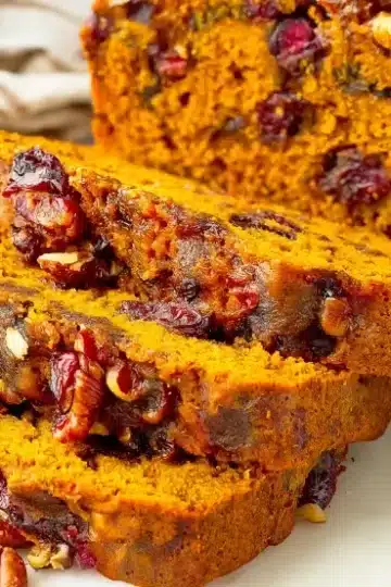 Three thick slices of cranberry pecan pumpkin bread in front of whole loaf, showing moist interior with cranberries and pecans visible, scattered nuts and cranberries on white surface