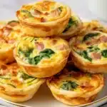 Close-up of golden baked mini quiches with spinach and diced ham stacked on a white plate.