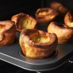 Multiple golden Yorkshire puddings with impressive rise and crispy ruffled edges in dark muffin tin, showing dramatic height and caramelized surfaces against dark background