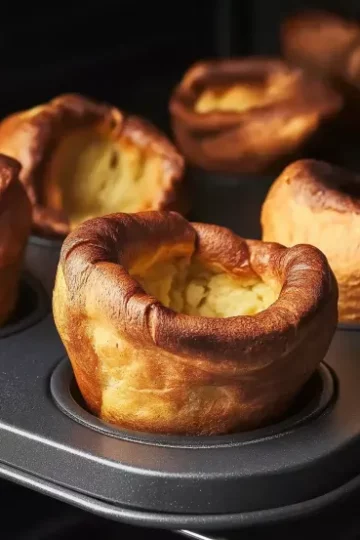 Multiple golden Yorkshire puddings with impressive rise and crispy ruffled edges in dark muffin tin, showing dramatic height and caramelized surfaces against dark background