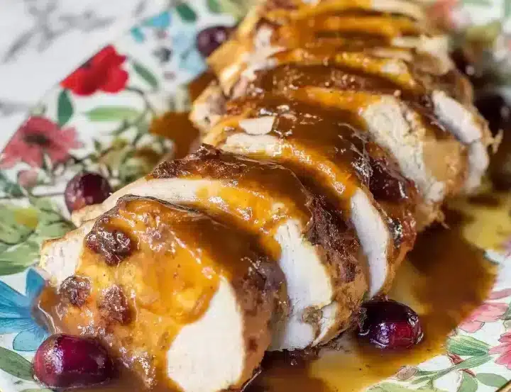 Tender sliced turkey breast on vintage floral plate with caramelized cranberry glaze drizzled between each slice, whole cranberries scattered around, golden sauce visible
