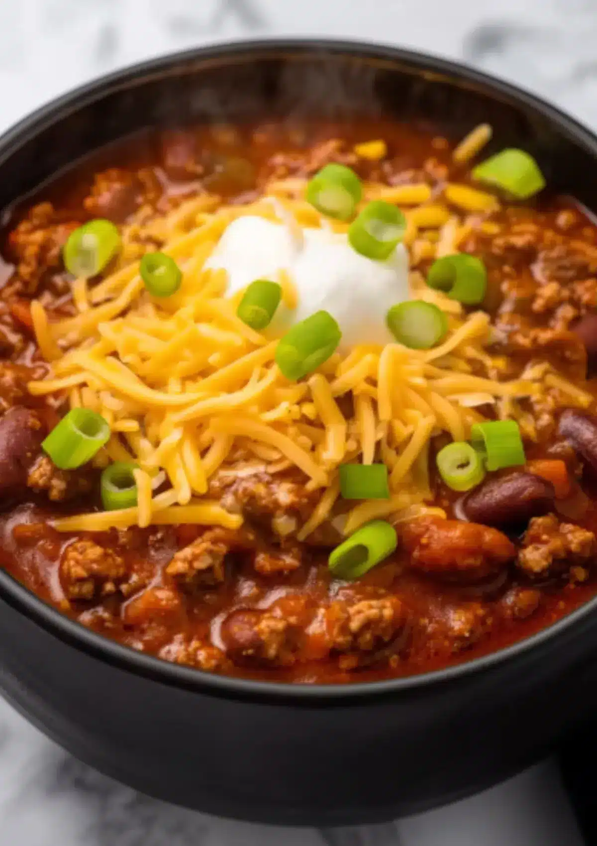 Close-up of hearty crock pot chili with ground beef and kidney beans in black bowl, topped with shredded cheddar cheese, sour cream, and fresh green onions