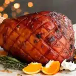 Whole glazed ham with cloth-wrapped bone handle, diamond scored pattern with cloves, garnished with fresh orange wedges and thyme sprigs on white parchment with warm Christmas lights bokeh