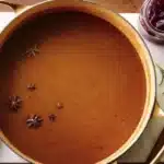 Overhead view of silky brown turkey gravy in gold-handled pot with floating star anise, fresh rosemary sprigs, wooden spoon and cranberry sauce jar on marble countertop, holiday meal prep