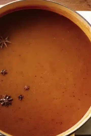 Overhead view of silky brown turkey gravy in gold-handled pot with floating star anise, fresh rosemary sprigs, wooden spoon and cranberry sauce jar on marble countertop, holiday meal prep