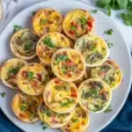 Overhead view of mini quiches on white plate showing variety of flavors with bacon, mushrooms, red bell peppers, fresh cilantro garnish, navy napkin and shredded cheese bowl