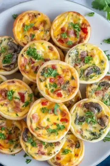 Overhead view of mini quiches on white plate showing variety of flavors with bacon, mushrooms, red bell peppers, fresh cilantro garnish, navy napkin and shredded cheese bowl