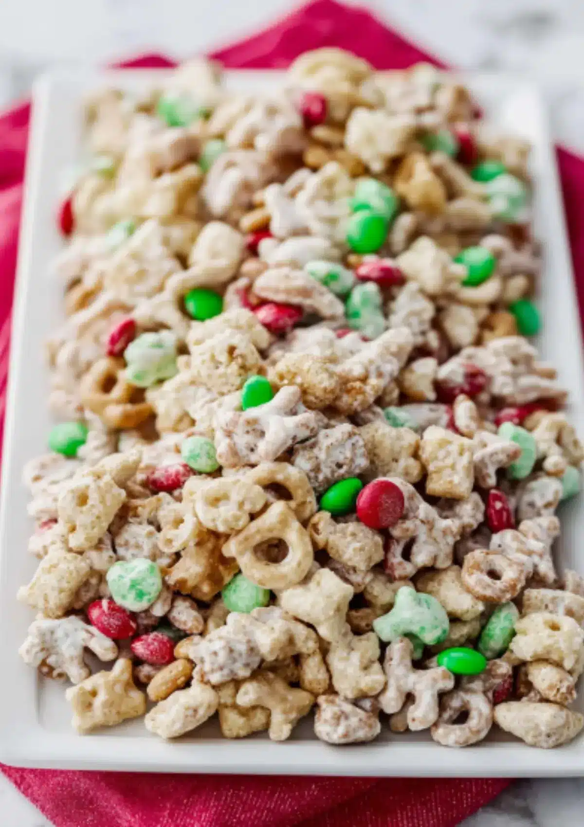 Close-up of reindeer chow featuring white chocolate coated Cheerios, Rice Chex, pretzels, peanuts and festive M&Ms on white rectangular platter with pink napkin