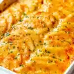 Angled view of three cheese scalloped potatoes in white baking dish showing golden brown bubbling cheese topping, layered potato slices with fresh parsley garnish on marble surface