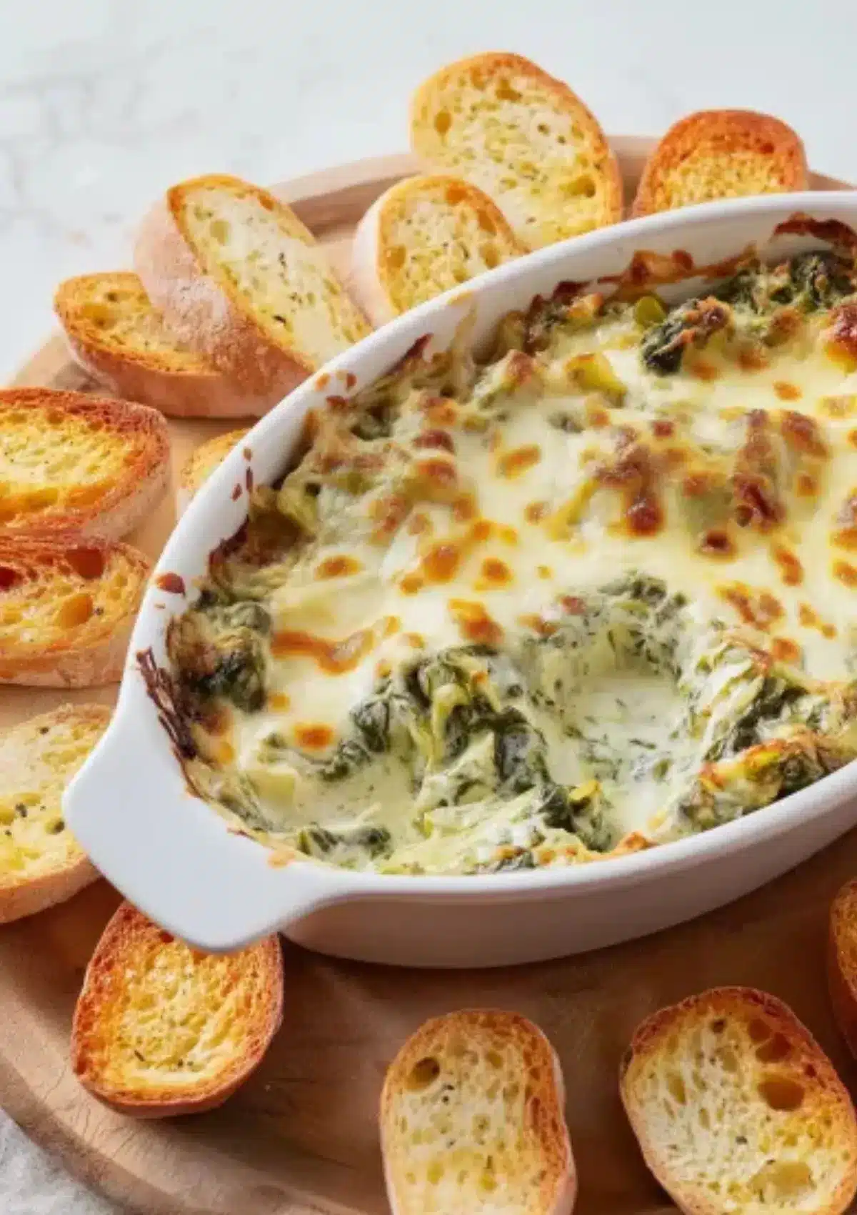 Close-up of hot bubbly spinach artichoke dip showing melted cheese, artichoke pieces, and spinach in white baking dish with golden brown garlic crostini on wooden board