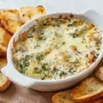Golden bubbly hot spinach artichoke dip in white ceramic baking dish surrounded by toasted garlic crostini on wooden serving board