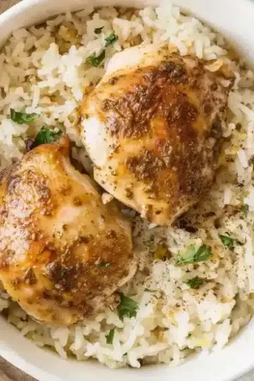 Two seasoned golden-brown chicken thighs with crispy herb crust served on bed of fluffy jasmine rice in white bowl, garnished with fresh parsley, photographed overhead on white marble countertop with tan linen napkin