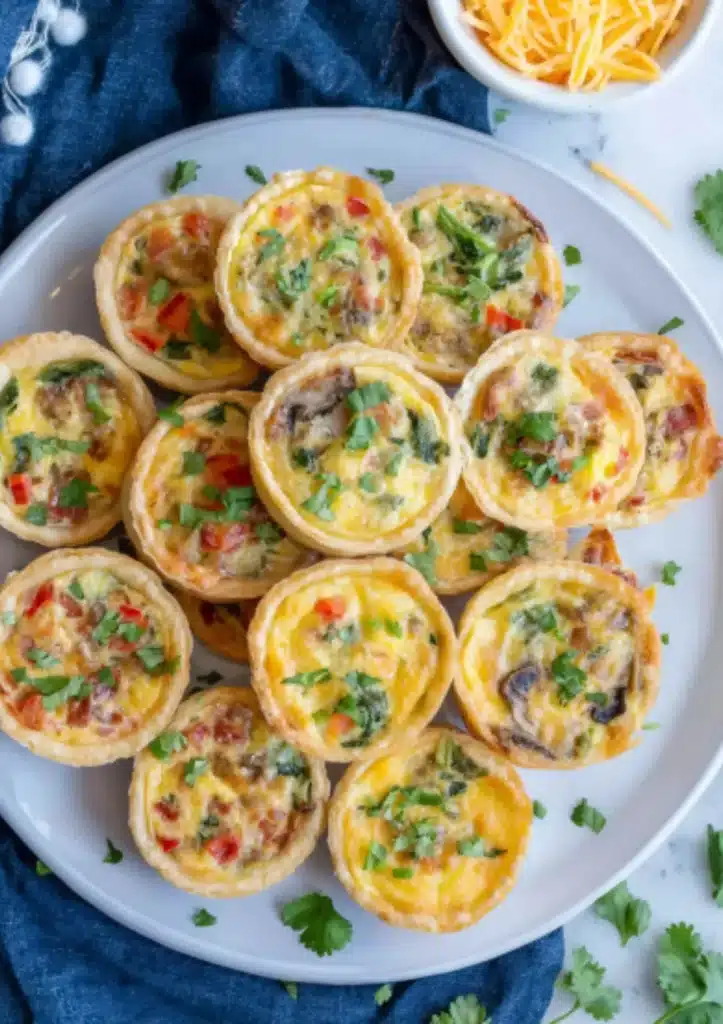 Overhead view of assorted mini quiches on white plate with colorful fillings including bacon, bell peppers, mushrooms, cheese and fresh cilantro garnish with navy napkin