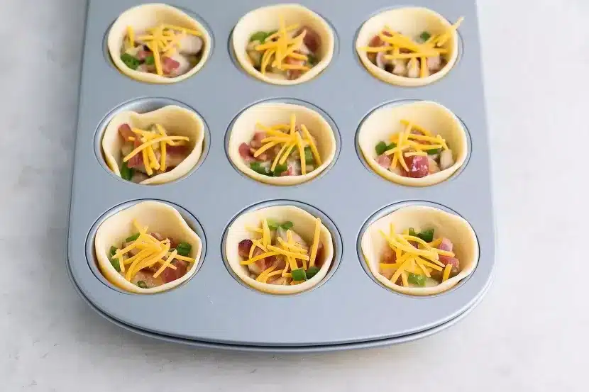Mini muffin tin with pie crust shells filled with diced bacon, bell peppers, mushrooms and shredded cheddar cheese before adding egg mixture