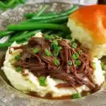 Complete Mississippi pot roast dinner featuring tender shredded beef over mashed potatoes with gravy, fresh green onion garnish, crisp green beans, and fluffy dinner roll on silver plate