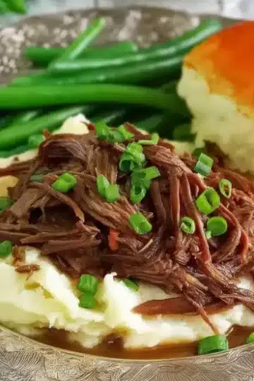 Complete Mississippi pot roast dinner featuring tender shredded beef over mashed potatoes with gravy, fresh green onion garnish, crisp green beans, and fluffy dinner roll on silver plate
