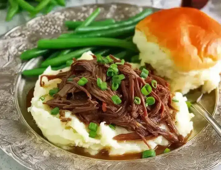 Complete Mississippi pot roast dinner featuring tender shredded beef over mashed potatoes with gravy, fresh green onion garnish, crisp green beans, and fluffy dinner roll on silver plate