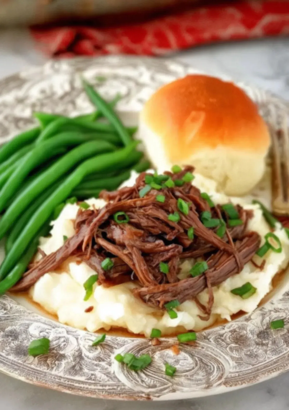 Shredded Mississippi pot roast piled on fluffy mashed potatoes with au jus gravy, garnished with fresh chopped green onions, served alongside bright green beans and golden dinner roll on vintage silver-patterned plate
