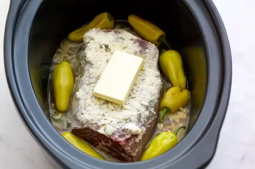 Raw chuck roast in slow cooker covered with ranch and onion soup mix seasoning, topped with full stick of butter and surrounded by 8 whole pepperoncini peppers ready to cook