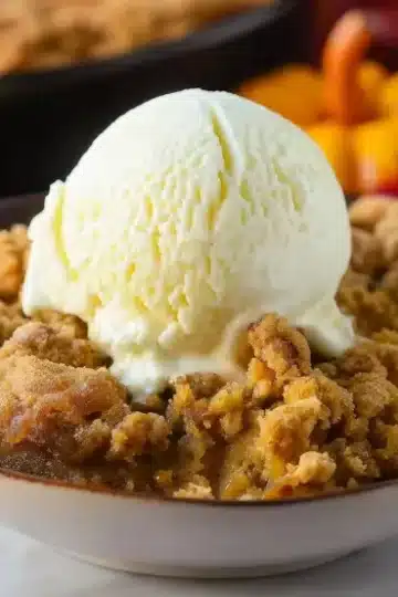 Close-up side view of pumpkin crisp in white bowl, vanilla ice cream scoop on golden cinnamon streusel topping, orange pumpkin filling visible, autumn leaves and pumpkins softly blurred in background