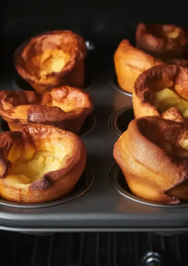 Six golden brown Yorkshire puddings with crispy edges and deep hollow centers in dark muffin tin against black background, showing dramatic rise and caramelization