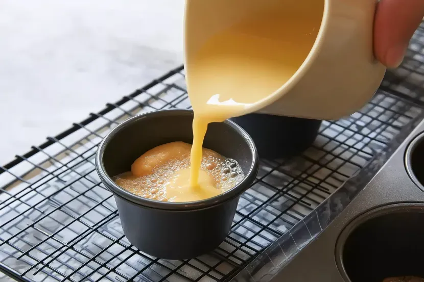 Pale yellow Yorkshire pudding batter being poured from cream ceramic jug into dark muffin tin on wire cooling rack over hot melted fat showing bubbles