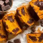 Flat lay overhead photo of sliced pumpkin chocolate chip bread on white marble with ceramic bowl of dark chocolate pieces showing moist orange crumb studded with melted chocolate