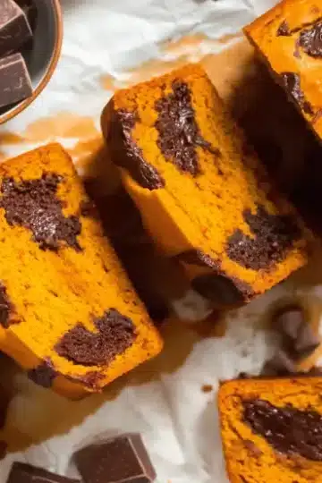 Flat lay overhead photo of sliced pumpkin chocolate chip bread on white marble with ceramic bowl of dark chocolate pieces showing moist orange crumb studded with melted chocolate