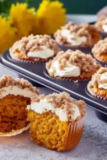 Pumpkin cream cheese crumble muffin cut open in foreground showing layers of golden pumpkin cake with thick white cream cheese filling and crumble topping, full muffin tin visible in background with yellow mum flowers