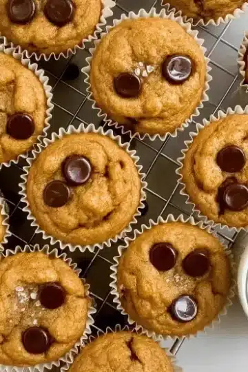 Overhead view of pumpkin protein muffins on black wire cooling rack with scattered dark chocolate chips, glass of milk, and orange striped kitchen towel on white marble surface