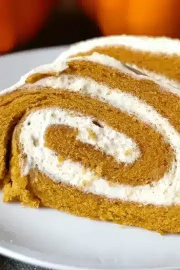 Extreme close-up of keto pumpkin roll slices showing perfect spiral swirl detail, golden spiced cake layers with bright white coffee cream filling, fine powdered sugar finish, artistic bokeh background with orange pumpkins