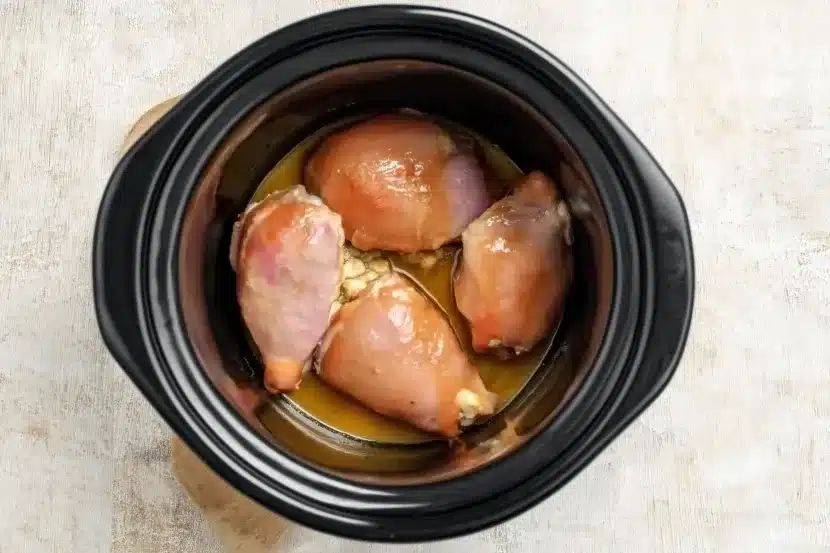Top-down view of four raw boneless chicken thighs arranged in black ceramic crock-pot with lemon juice mixture and visible minced garlic, ready for slow cooking on light textured surface