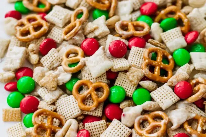 Macro close-up of reindeer chow showing detailed texture of white chocolate coating on Rice Chex squares, pretzel twists, and colorful red and green M&Ms
