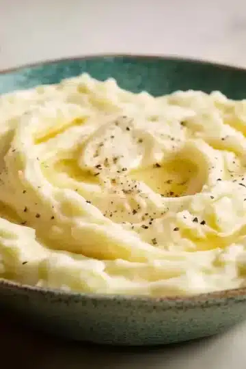 Perfectly whipped mashed potatoes served in handcrafted teal ceramic bowl with butter drizzle and pepper, butter dish and olive oil visible in background