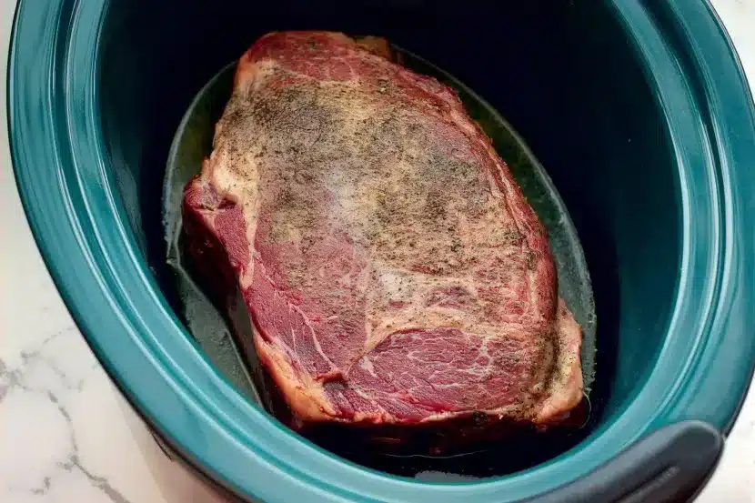 Raw marbled chuck roast seasoned with salt, pepper and herbs in teal slow cooker insert, overhead view showing preparation stage of Mississippi pot roast recipe